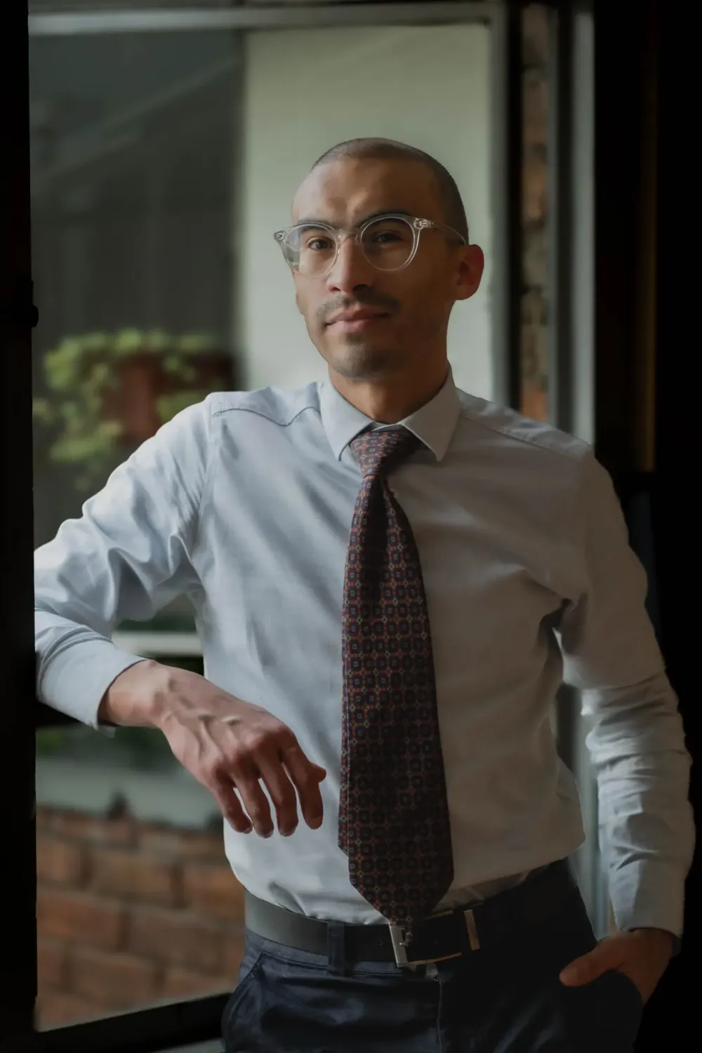 Hombre en camisa de vestir y corbata estampada posando junto a una ventana
