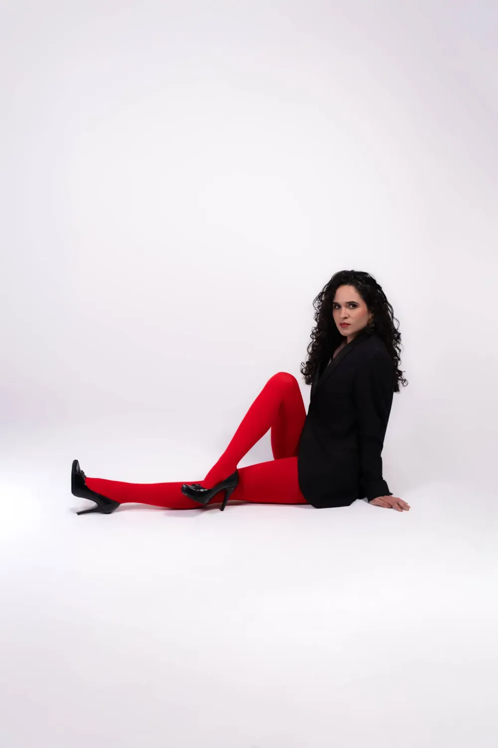 Mujer en blazer negro y medias rojas sentada en piso blanco de estudio